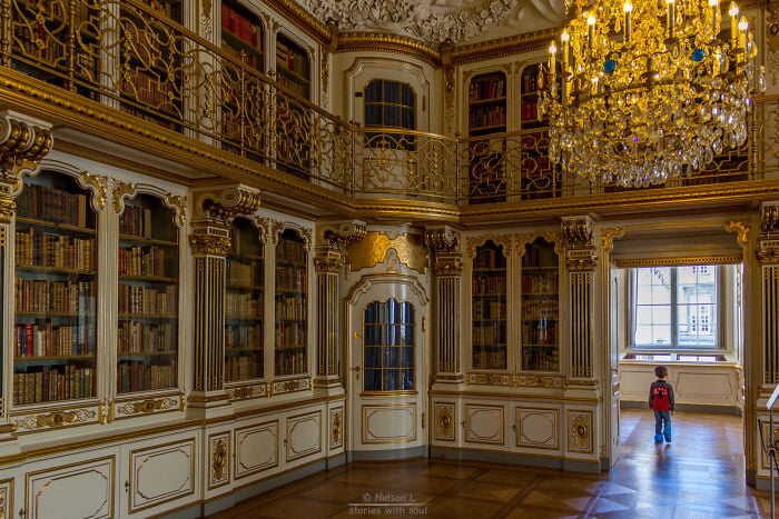 Interior grandioso de biblioteca com detalhes dourados ornamentados, estantes altas e uma crian&ccedil;a explorando um cen&aacute;rio m&aacute;gico de livraria.