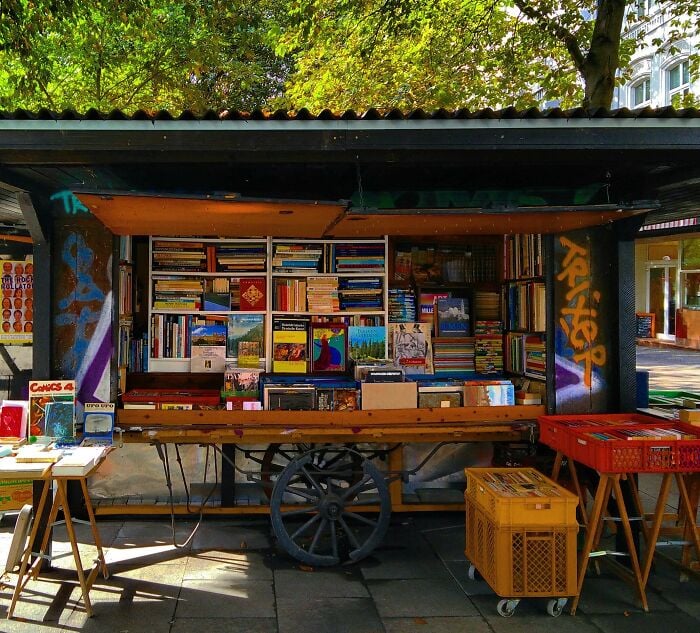 Livraria aconchegante ao ar livre com prateleiras cheias de livros, cercada por vegeta&ccedil;&atilde;o, exibindo bibliotecas e livrarias m&aacute;gicas.
