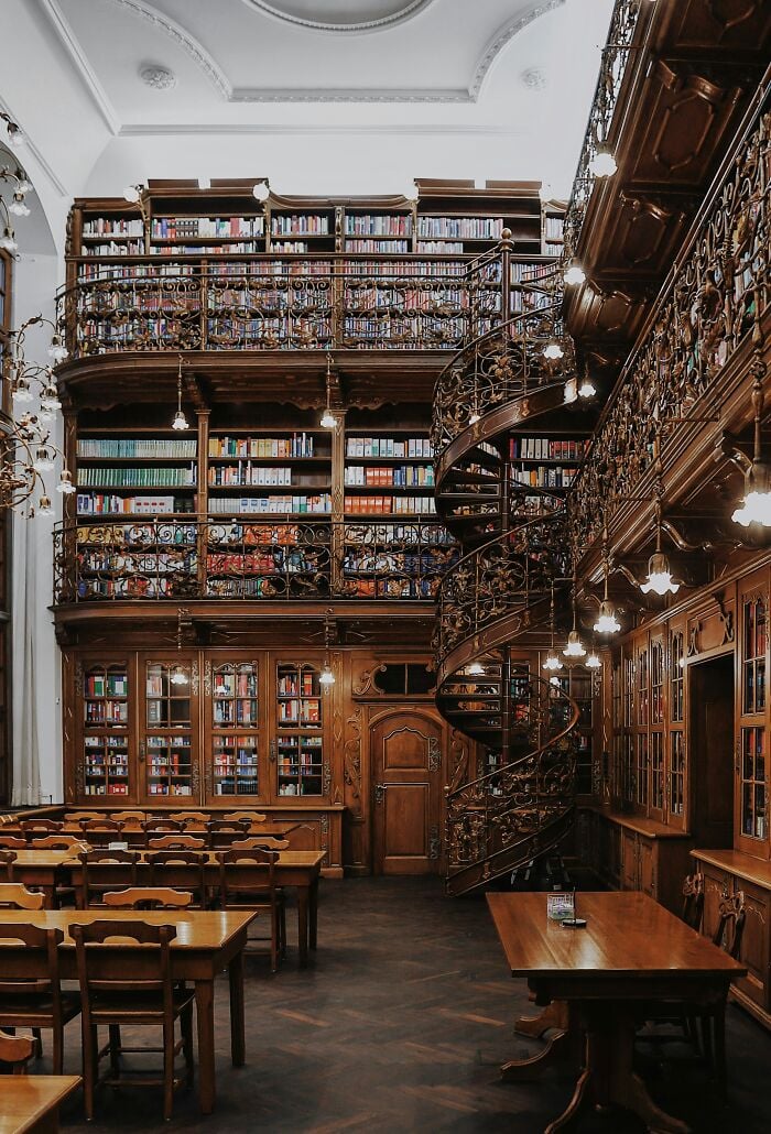 Interior grandioso de biblioteca com prateleiras de madeira, escadaria em espiral e aconchegantes mesas de leitura cheias de livros de todo o mundo.