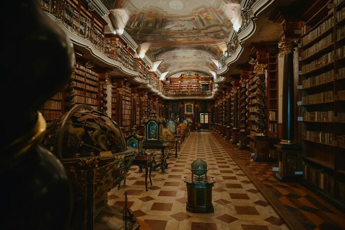 Interior grandioso de uma biblioteca com globos antigos, colunas ornamentadas e fileiras de livros antigos em prateleiras de madeira.