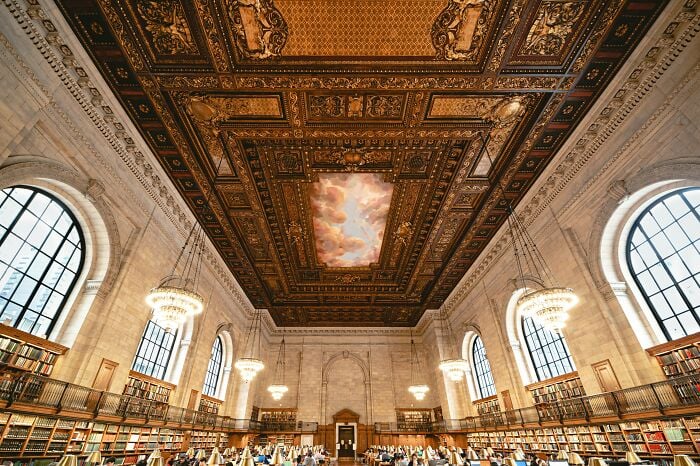 Interno di una grandiosa biblioteca con soffitto ornato, grandi finestre ad arco, lampadari e file di scaffali pieni di libri.