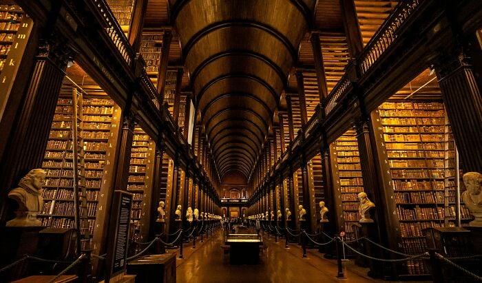 Interior de biblioteca grandioso e acolhedor com estantes imponentes, arcos de madeira e bustos de mármore alinhando um grande corredor.